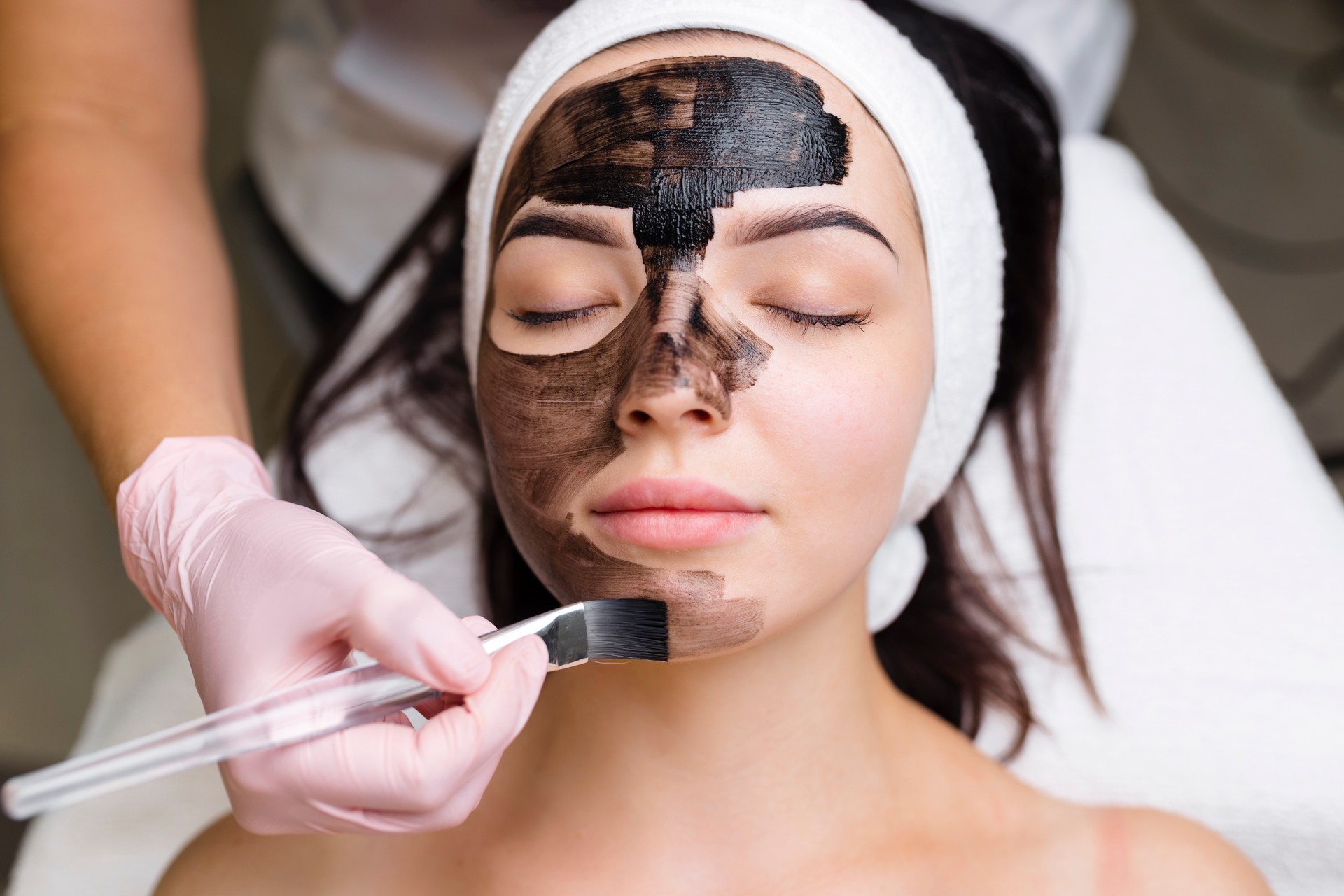 cosmetologist applying black mask on pretty woman face wearing black gloves, gorgeous woman in spa having facial procedures